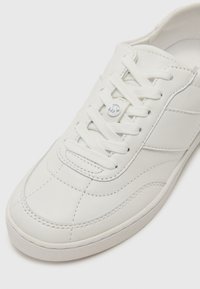 White leather sneaker featuring a rounded toe, textured surface, flat laces, and a subtle logo detail on the side.