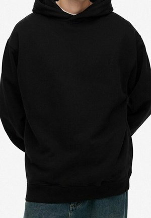 Person trägt einen schlichten schwarzen Kapuzenpullover mit langen Ärmeln und blaue Jeans, steht mit den Händen hinter dem Rücken.
