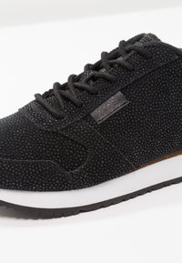 Baskets en daim noir avec des points texturés, lacets noirs, semelle blanche et une petite étiquette en tissu sur le côté, sur fond blanc.