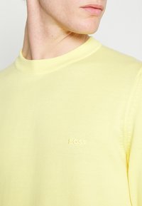 Nahaufnahme einer Person, die einen hellgelben Rundhals-Pullover mit dem Wort "BOSS" auf der Brust trägt.
