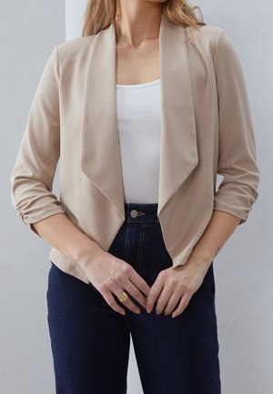 Femme portant un blazer beige à ouverture frontale, un haut blanc et un jean bleu foncé avec des manches froncées et une bague en or à la main gauche.