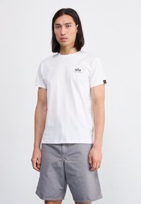 Weißes Baumwoll-T-Shirt mit Rundhalsausschnitt, kurzen Ärmeln, kleinem schwarzem Logo auf der Brust, getragen mit hellgrauen Shorts. Einfaches und lässiges Design.