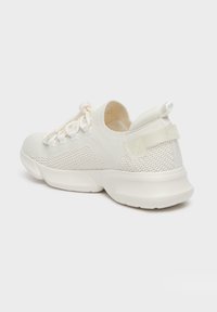 Zapatilla deportiva color crema con parte superior de malla texturada, cordones elásticos y suela acolchada con agarre de goma. La parte trasera cuenta con una pestaña de agarre.