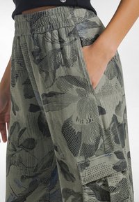 Pantalones cargo verdes con un estampado floral negro, cintura elástica, bolsillo lateral y tela texturizada con patrones y acentos variados.