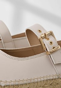 Helle beigefarbene Lederschuhe mit Jutesohle, die eine goldfarbene Schnalle, kontrastierende Nähte und ein offenes Rückendesign aufweisen.