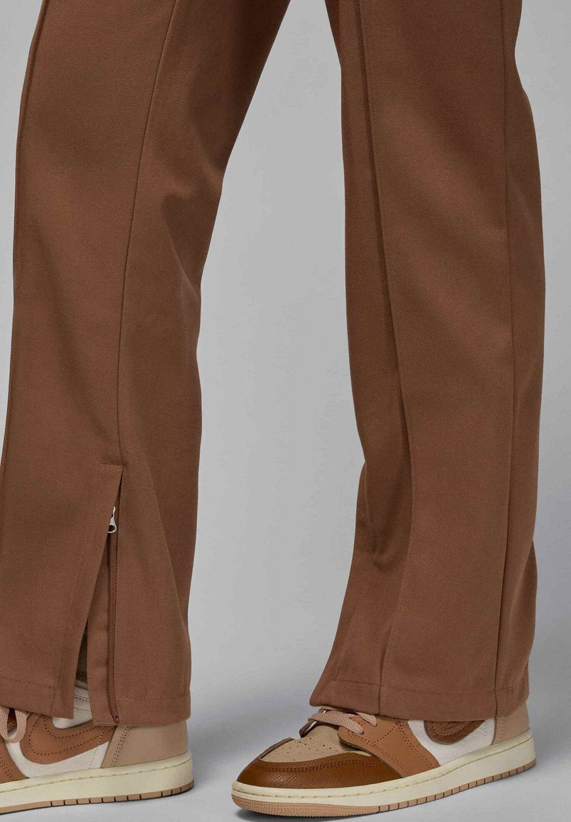 Pantaloni dritti marroni con dettaglio della zip laterale indossati con scarpe da ginnastica marroni e beige con lacci su uno sfondo grigio.