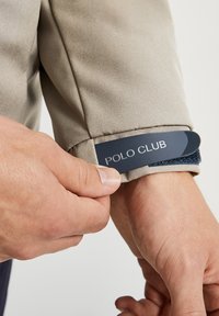 Ruka upravuje béžový rukáv saka se zapínáním na modrý suchý zip označeným "POLO CLUB" na jednobarevném pozadí.