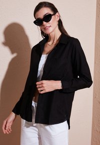 Black long-sleeve button-up shirt with a pocket, soft fabric, slightly relaxed fit, worn over a white top and paired with white pants.