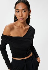 Sort top med lange ærmer, en off-shoulder design, rynket tekstur og en tætsiddende silhuet. Bæres med højtaljede bukser.
