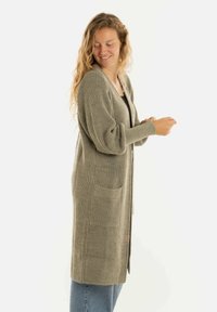 Vrouw met een glimlach, gekleed in een lange groene gebreide cardigan met zakken over een zwart topje en blauwe spijkerbroek, staand tegen een witte achtergrond.