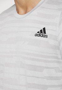 Camiseta deportiva gris claro con un patrón texturizado, cuello redondo y un logo negro de Adidas en el pecho.