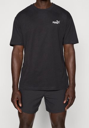 Mężczyzna w czarnej koszulce Puma i czarnych szortach, stojący na tle jednolitego jasnego tła, z rękami swobodnie opuszczonymi wzdłuż ciała.