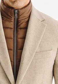 Mężczyzna w beżowej żakietowej kurtce z prążkowanej tkaniny, noszący brązową pikowaną kurtkę z zapięciem aż do szyi.