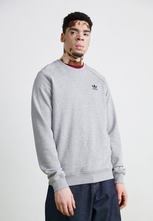 Grå sweatshirt med rund hals og ribbede manchetter, der har et lille sort logo på brystet. Kombineret med mørke jeans.