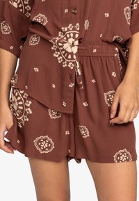 Rompers à motifs floraux bruns avec manches courtes, taille cintrée et fermeture à boutons. Le tissu semble léger avec une coupe décontractée.