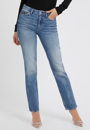 Vrouw draagt lichtblauwe rechtlopende spijkerbroek, zwart lange mouwen top, en met strass-stenen versierde hoge hakken sandalen, hand in de zak.