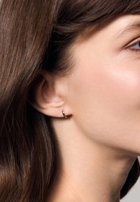 Złoty kolczyk w kształcie koła noszony w lewym uchu, o gładkim wykończeniu z delikatnym zakrzywieniem, zestawiony z średniobrown włosami i jasną karnacją.