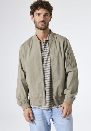 Olivgrüne Bomberjacke mit gerippten Bündchen, Reißverschluss und Seitentaschen, darüber ein gestreiftes Hemd, kombiniert mit hellblauen Jeans.