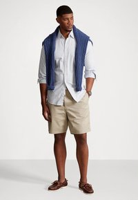 Kasdienis aprangos komplektas su mėlynu džemperiu su pintomis kilpomis, užmestu ant baltos dryžuotos palaidinės, chaki spalvos šortais ir rudos odos valtimi batais.