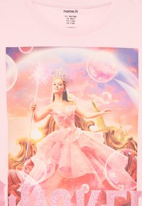 Šviesiai rožinė medvilninė marškinėliai su spalvinga grafika, vaizduojančia princesę rožiniame suknelėje, apsuptą burbulų, su blizgučiais užrašu "WICKED".