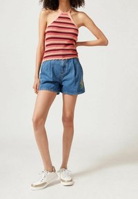 Donna con un top a maniche scoperte a righe rosa e rosse, pantaloni denim blu con ricami floreali e sneakers bianche con dettagli dorati.