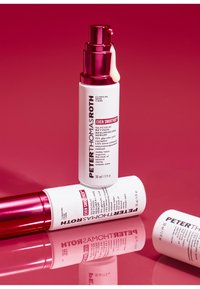 Twee witte serumflessen met roze accenten, voorzien van pompdispensers. Etiketten bevatten de productnaam en ingrediënten. Reflecterend oppervlak eronder.