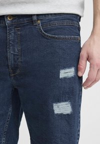 Sötétkék farmernadrág, megviselt foltokkal. Klasszikus öt zsebes kialakítással és látható gombos, cipzáras záródással rendelkezik.