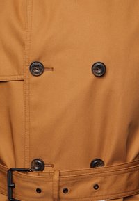 Cappotto doppiopetto in tessuto beige con una superficie liscia. Presenta bottoni scuri e una cintura coordinata con una fibbia in metallo nero.