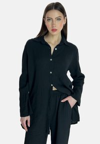 Camisa negra de botones, hecha de tejido texturizado, con cuello, mangas largas y un corte holgado, combinada con pantalones a juego.