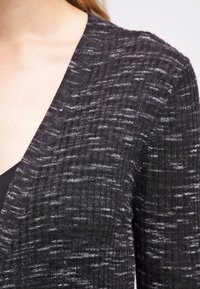 Lähikuva henkilöstä, joka on pukeutunut tummanharmaaseen, ribbib neulepuseroon, jossa on valkoisia pilkkuja mustan paidan päällä.