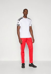 Balta sporta T-krekls ar melnām svītrām, kopā ar sarkaniem sporta biksēm ar baltām sānu svītrām; modelis valkā melnus sporta apavus.