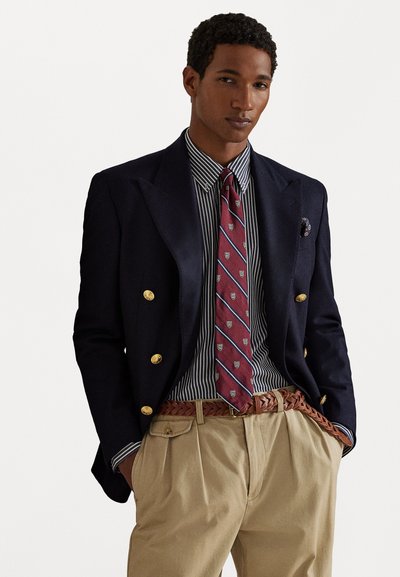 Ναυτικό σακάκι με χρυσά κουμπιά πάνω από πουκάμισο με ρίγες. Βουργουνδίας γραβάτα με σχέδια, καλα�ίσθητο παντελόνι καμισό, και πλεκτή καφέ δερμάτινη ζώνη.