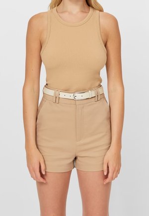 Woman wearing a sleeveless beige ribbed top tucked into high-waisted beige shorts with a light-colored belt.