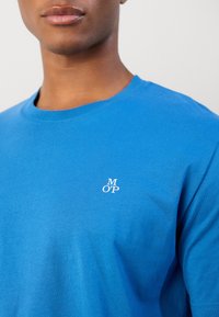 T-shirt de algodão azul com decote redondo, apresentando um logotipo branco "MOP" no lado esquerdo do peito. Textura suave, design casual.