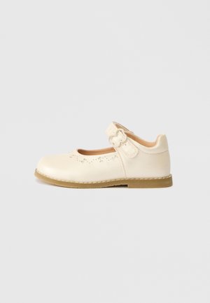Chaussure Mary Jane écrue avec bride à boucle, motif floral perforé près des orteils et semelle plate marron clair sur fond uni.