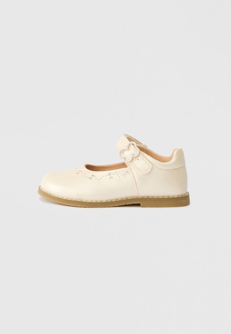 Chaussure Mary Jane écrue avec bride à boucle, motif floral perforé près des orteils et semelle plate marron clair sur fond uni.