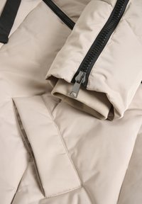 Beige pufferjack met een gestructureerde buitenkant, zwarte rits en een ribbelmanchet zichtbaar bij de mouwopening. Heeft een verborgen zak.