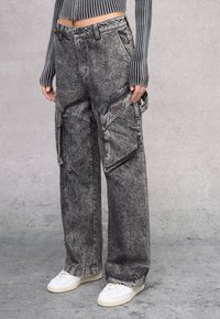 Baggy, high-waisted cargo jeans van vervaagd zwart denim met contrasterende stiksels, grote zijzakken en een rauwe zoom. Gecombineerd met witte sneakers.