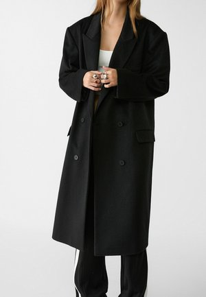 Personne portant un long manteau noir à double boutonnage par-dessus un haut blanc et un pantalon noir, mains jointes montrant plusieurs bagues.