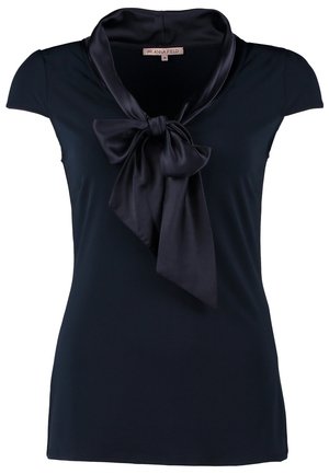 Femme portant un chemisier bleu marine à manches courtes avec un nœud en satin, un jean bleu, des bottines noires, tenant une pochette foncée, marchant vers l'avant.