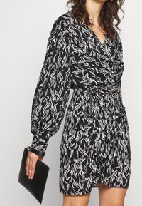 Robe à motifs noir et blanc avec de longues manches bouffantes, un col en V et une taille nouée. Le mannequin tient une pochette noire.