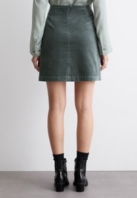 Falda de pana verde con cintura ajustada, cremallera lateral y bordes dobladillados. Llevada con botas negras hasta el tobillo y calcetines oscuros, vista desde la parte posterior.