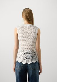 Hvit strikket ermeløs topp med åpen blondermønster, bølget kant, og en litt lengre bakdel, kombinert med løse denimjeans.