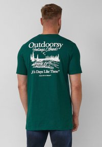 Man in een donkergroen T-shirt met witte buiten-natuurafbeelding en tekst op de rug, gecombineerd met donkerblauwe jeans.