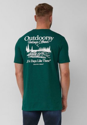 Mann trägt ein dunkelgrünes T-Shirt mit weißem Naturmotiv und Text auf dem Rücken, kombiniert mit dunkelblauen Jeans.