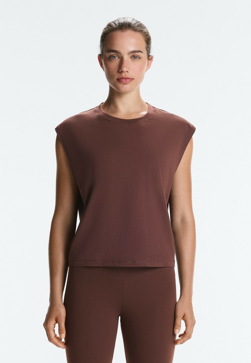 Femme aux cheveux tirés en arrière portant un haut marron sans manches et un pantalon marron ajusté assorti, debout devant un fond clair uni.