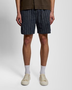 Man met donkerblauwe gestreepte korte broek, witte sokken en crème kleurige veter sneakers, staand tegen een effen lichte achtergrond.