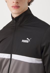Črna in siva športna jakna z visokim ovratnikom, belo vodoravno črto in belim logotipom Puma na prsih. Gladka tekstura tkanine.