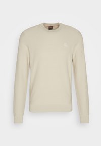 Suéter de punto color beige claro con cuello redondo, puños y dobladillo acanalados, y un pequeño parche de marca en el pecho. Detalle de tejido texturizado visible.