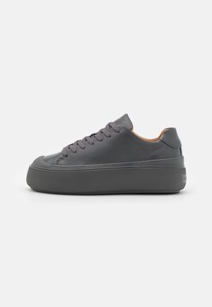 Zapatilla deportiva baja de color gris oscuro con suela gruesa, puntera redonda y cordones a juego en gris, mostrada desde el lado exterior sobre un fondo blanco.